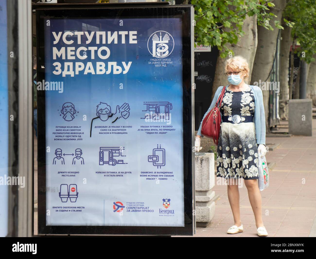 Belgrade, Serbie - 15 mai 2020 : une femme blonde mature portant un masque chirurgical et des gants debout seule à l'arrêt d'autobus avec des directives sur le coronavirus Banque D'Images