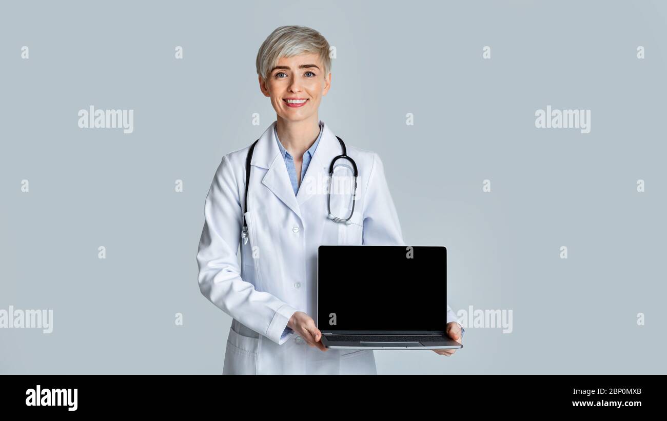 Une femme souriante, médecin manteau blanc, montre un ordinateur portable avec un écran vierge. Banque D'Images