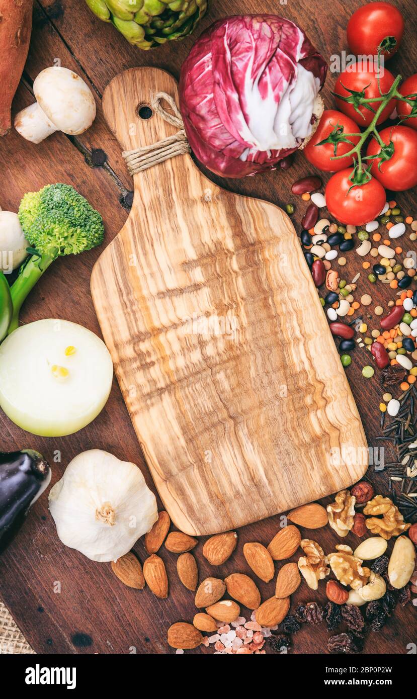 Planche à découper en bois vide entourée de légumes frais et non cuits et de noix. Modèle d'aliments sains, espace de copie, photo verticale, vue du dessus. Banque D'Images