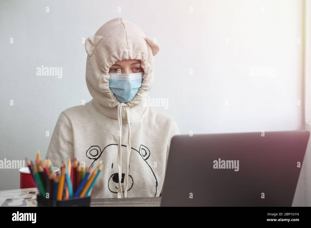 Jeune fille portant un masque et un drôle de sweat à capuche regardant s'ennuyer devant un ordinateur portable à la maison. Travailler à domicile pendant le concept de quarantaine covid Banque D'Images
