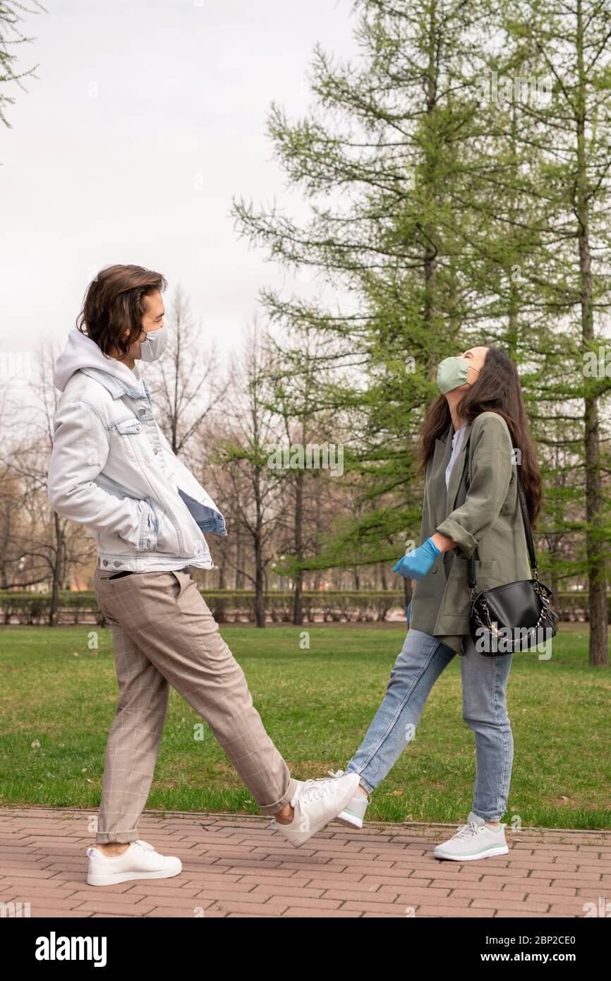 Jeune couple gai en vêtements décontractés et masques protecteurs debout dans le parc au milieu des arbres verts et des pelouses et profiter de la chaude journée d'été Banque D'Images