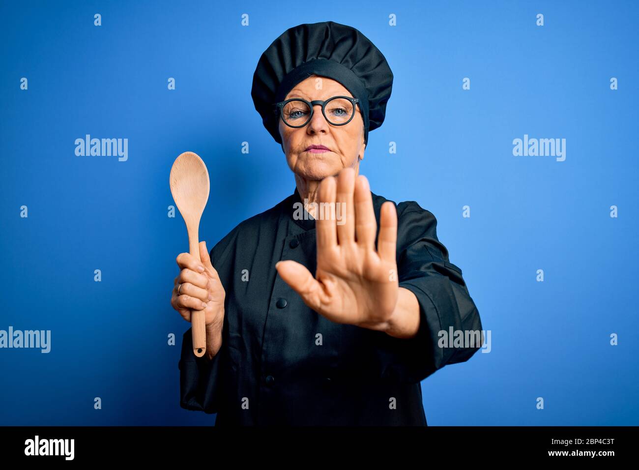 Senior belle femme de chef à cheveux gris portant l'uniforme de cuisinière et le chapeau tenant une cuillère en bois à main ouverte faisant signe d'arrêt avec sérieux et confiant Banque D'Images