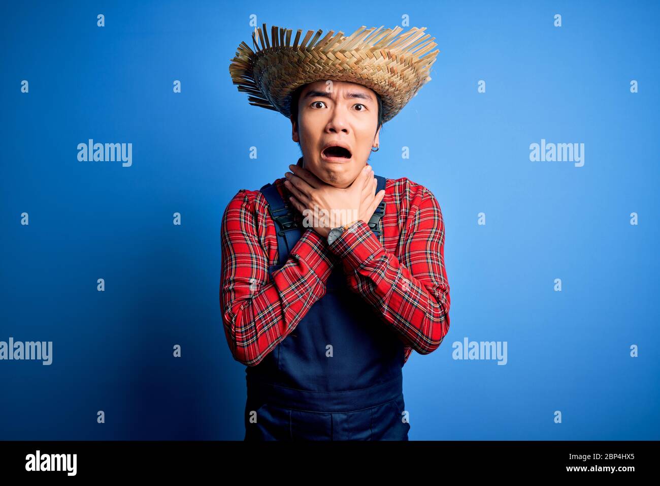 Un jeune fermier chinois charmant portant un tablier et un chapeau de  paille sur fond bleu criant et suffoquer parce que la cornière douloureuse.  Problème de santé. Comme Photo Stock - Alamy