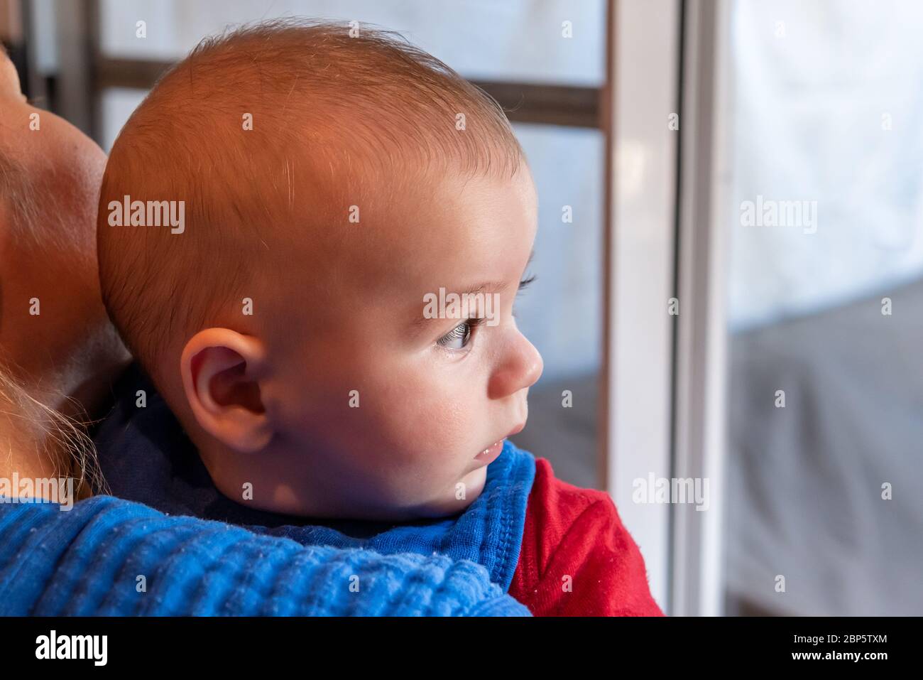 Mignon 6 mois petit garçon avec la curiosité expression sur son visage dans ses bras de mères Banque D'Images