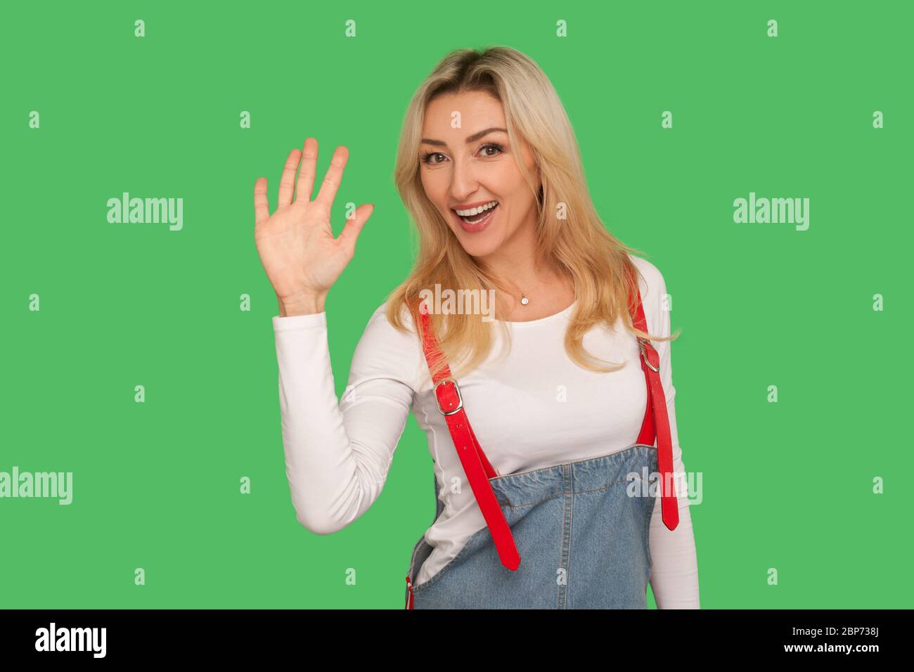 Bonjour, bienvenue ! Portrait de charmante femme blonde adulte aimable dans des combinaisons en denim élégantes levant la paume à la vague hi, salutation avec amical accueillant sm Banque D'Images