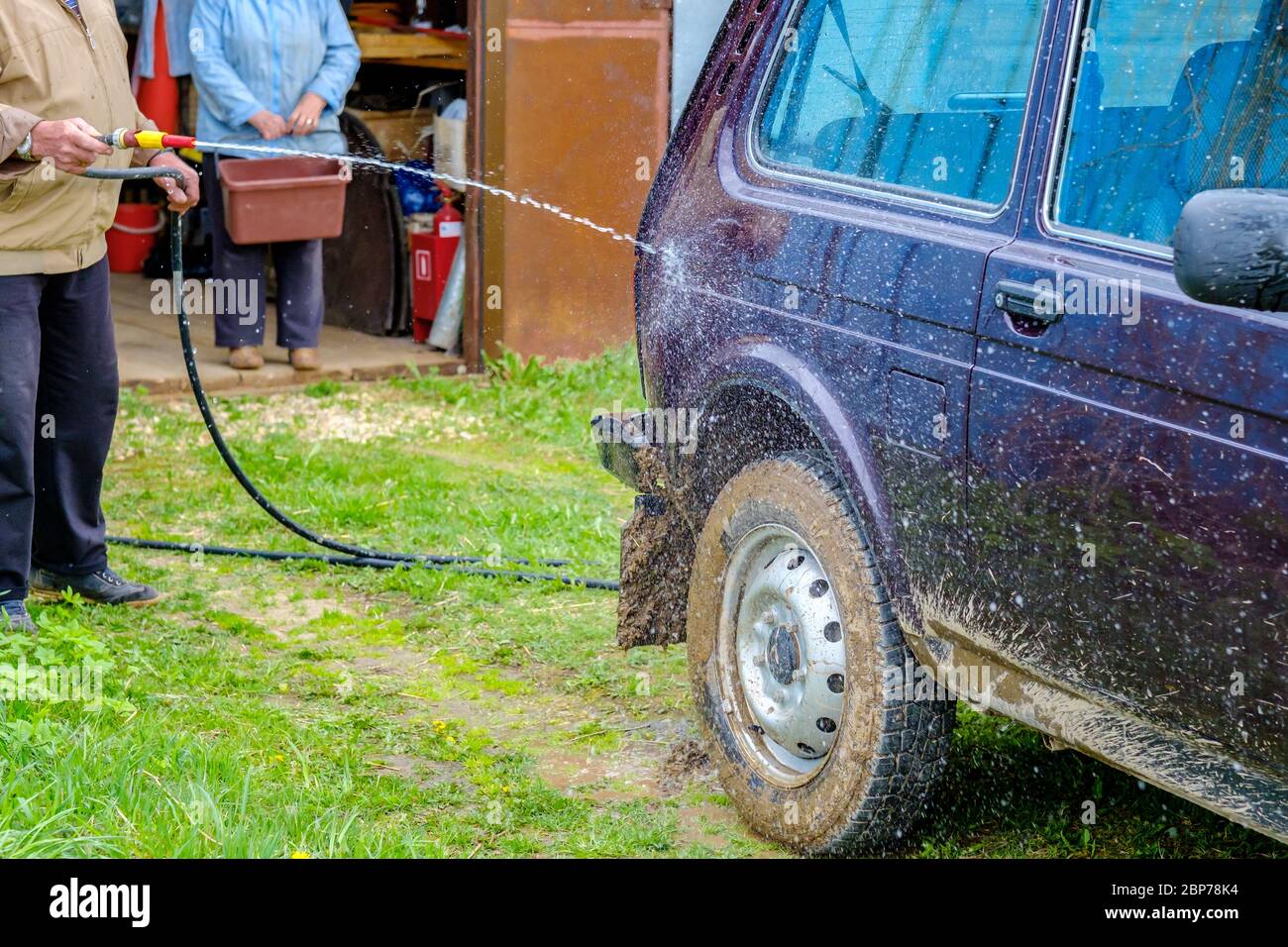 Un homme âgé aux cheveux gris lave une voiture à l'extérieur avec un tuyau  d'arrosage. Un puissant jet d'eau évacue la saleté du corps violet de la  voiture, du verre Photo Stock -