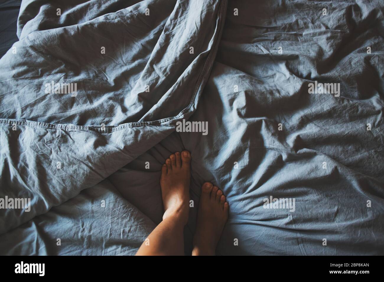 Vue de dessus lit froissé le matin après le sommeil. Draps en coton gris et couvertures sur le lit. Jambes de femme Banque D'Images
