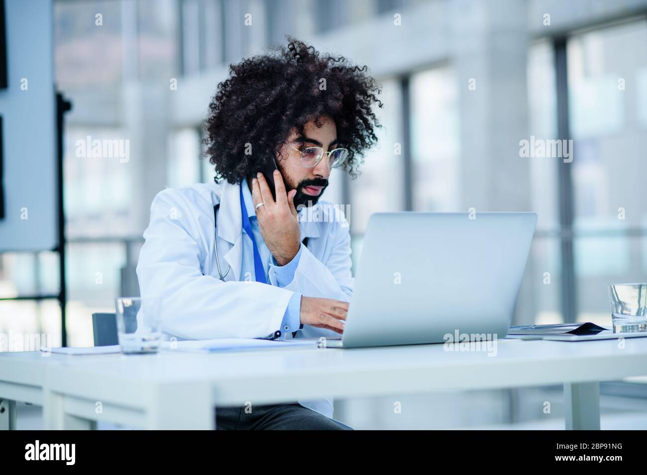 Portrait d'un médecin de sexe masculin assis à l'hôpital, à l'aide d'un ordinateur portable et d'un smartphone. Banque D'Images