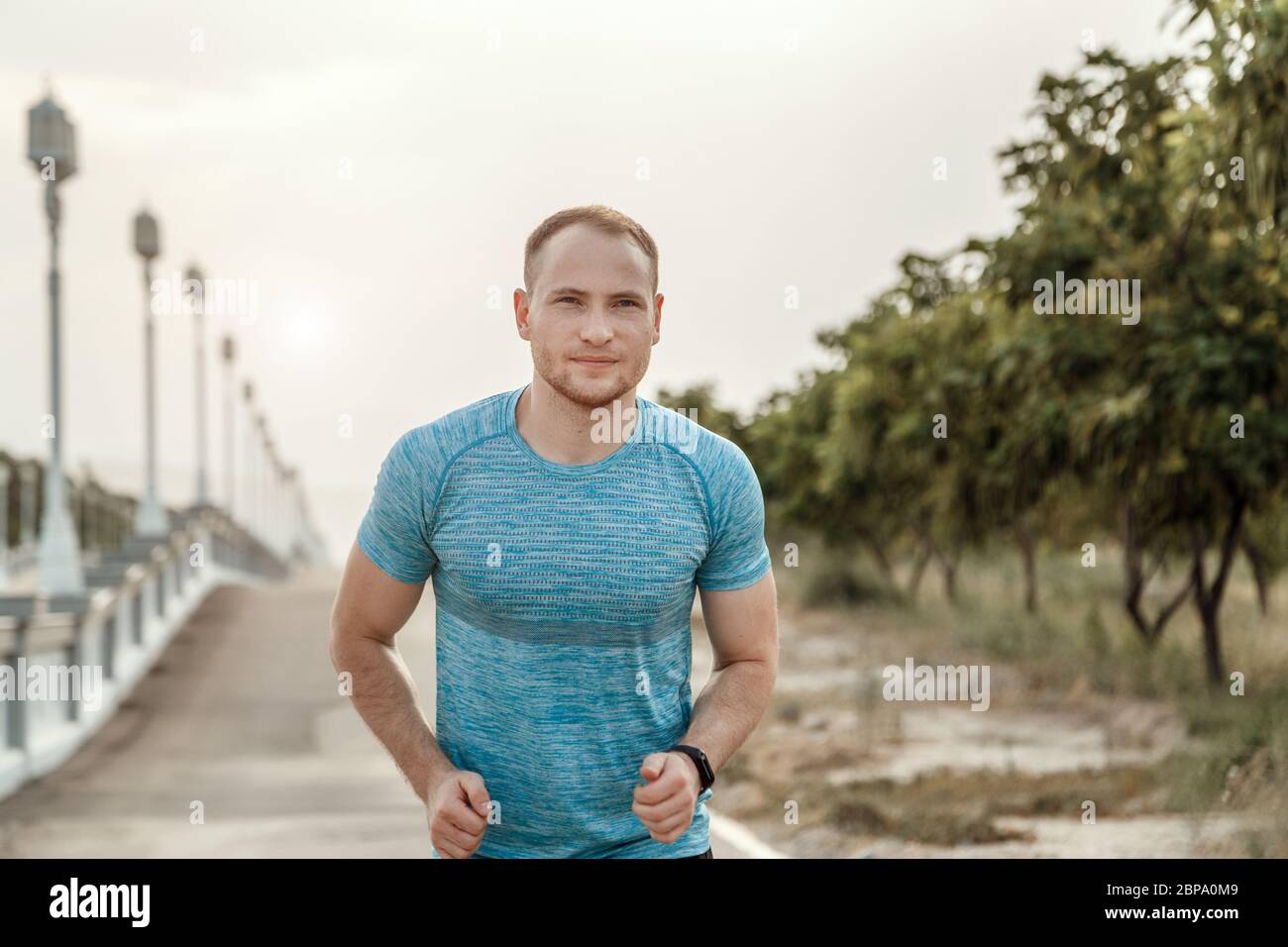 Portrait of Caucasian guy dans un tee-shirt bleu et un short noir qui s'entraîne et s'exécute sur la piste asphaltée pendant le coucher du soleil Banque D'Images