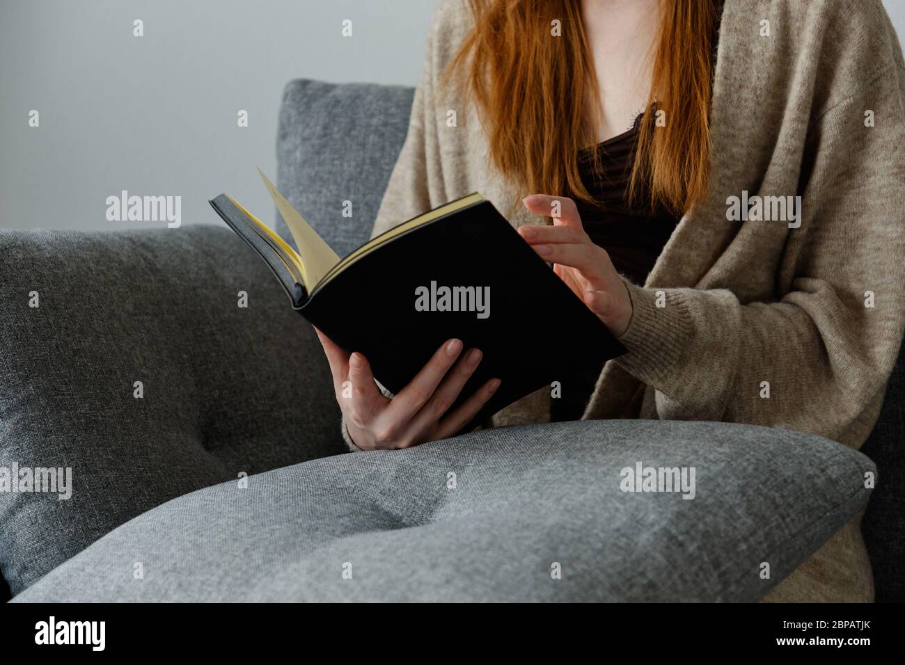 Un livre noir dans les mains d'une fille à cheveux rouges assise sur un canapé. Banque D'Images