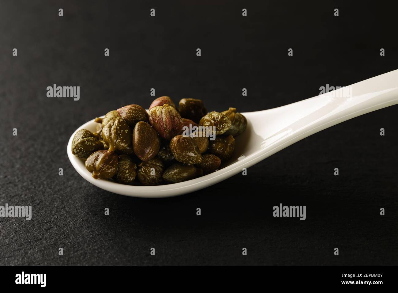 Câpres marinés dans une cuillère en porcelaine chinoise blanche sur une planche de service en pierre noire. Sauces et assaisonnements pour la viande, le poisson et les légumes. Vue avant. Banque D'Images