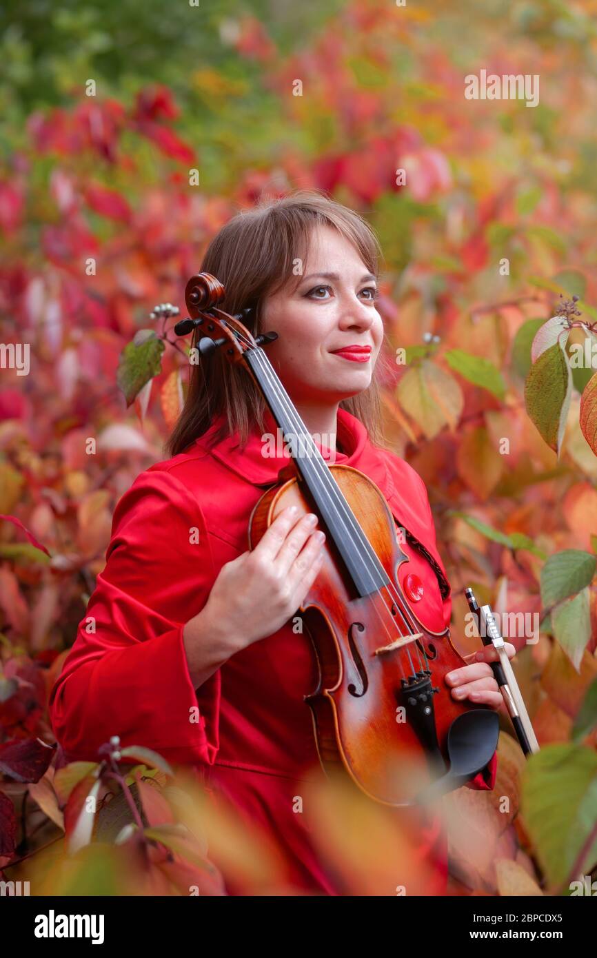 jeune belle femme en manteau rouge tenant le violon dans les mains souriant et se cachant parmi les feuilles rouges et vertes de couleur d'automne dans la forêt ou le parc d'automne Banque D'Images
