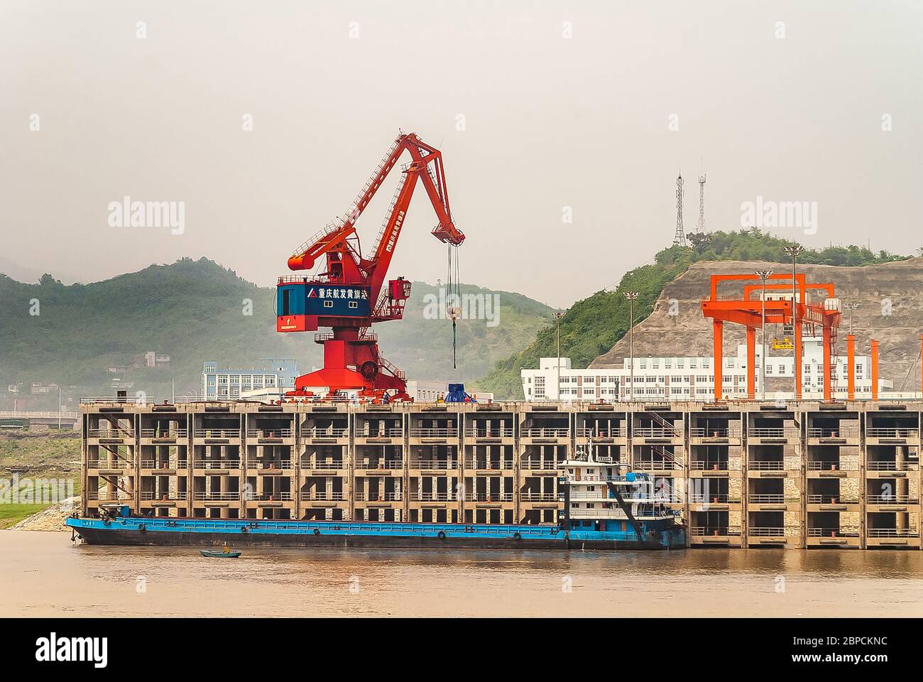 Huangqikou, Chongqing, Chine - 8 mai 2010 : fleuve Yangtze. La grue rouge et bleue décharge la barge bleue au quai à plusieurs niveaux du port. Colline verte sous smog sk Banque D'Images