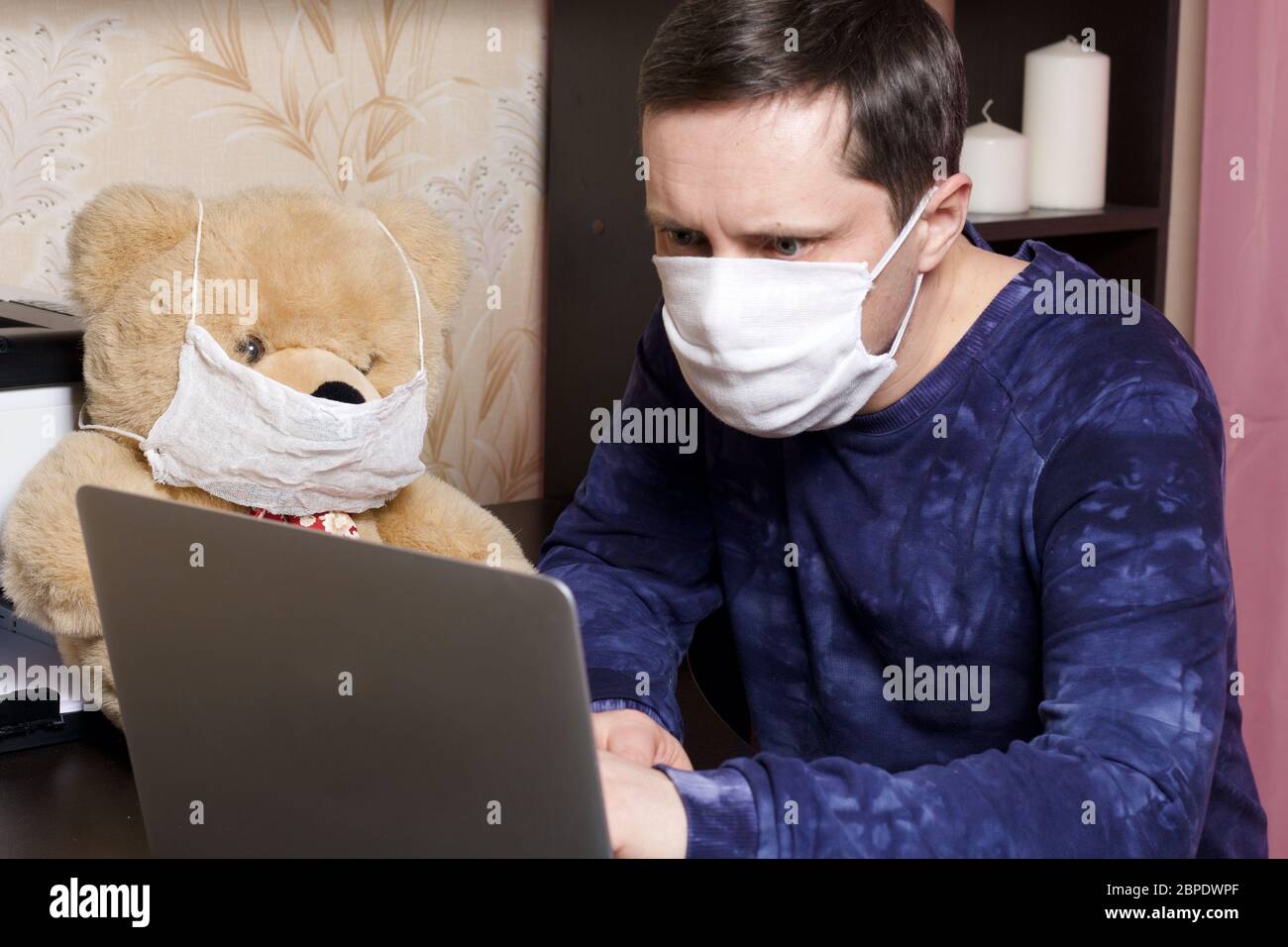 Un homme dans un masque de gaze de protection travaille derrière un ordinateur portable dans des conditions d'auto-isolation. À proximité, vous trouverez un jouet. Porter un masque de gaze de protection. Banque D'Images