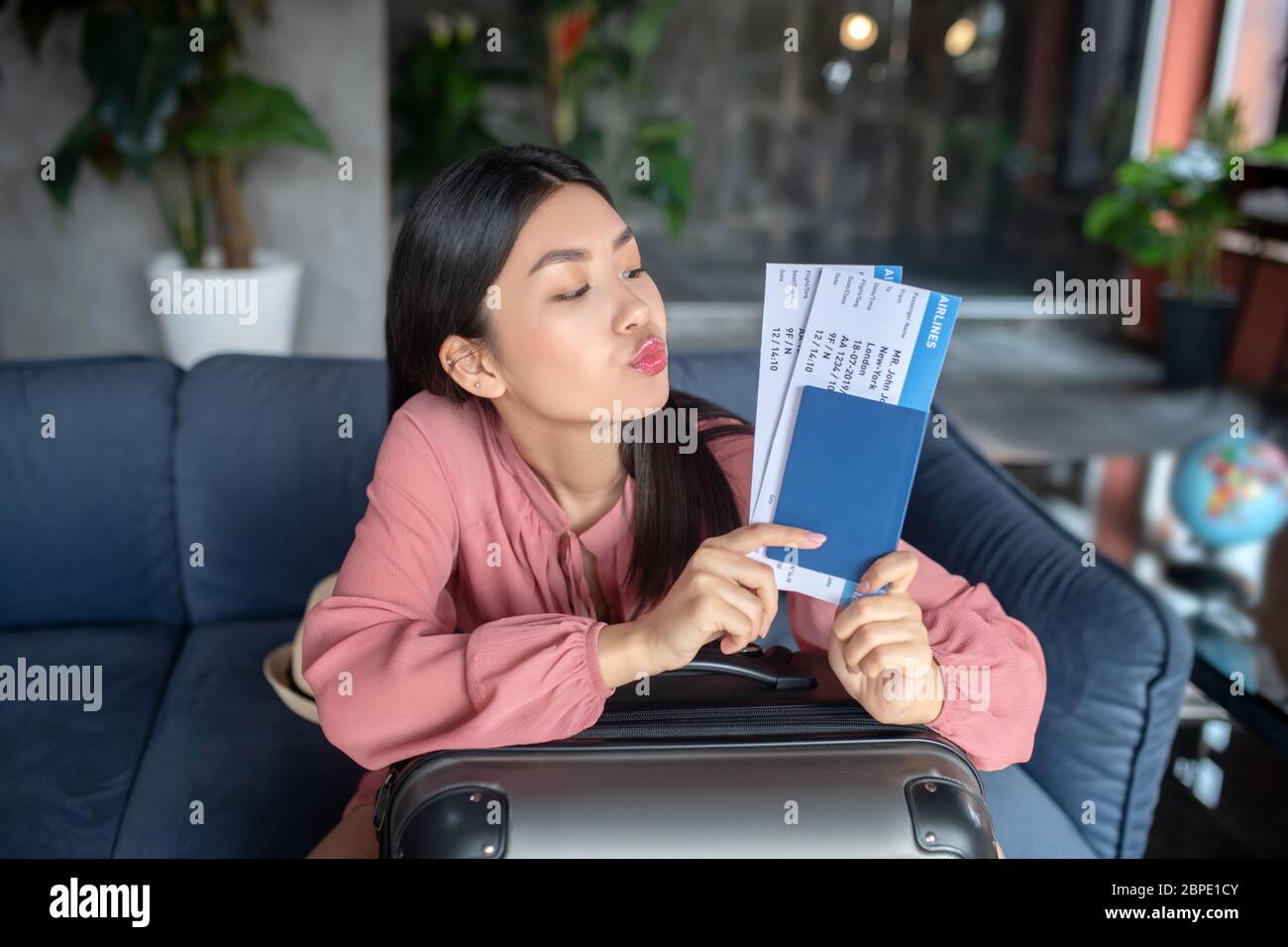 Heureuse femme aux cheveux foncés assise sur un canapé, tenant des billets d'avion, ses lèvres donnant baiser Banque D'Images