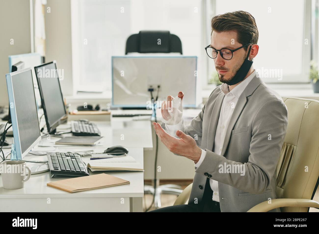 Jeune homme sérieux et beau avec masque sur le menton pulvérisant un antiseptique sur les mains tout en travaillant seul dans un bureau vide pendant le coronavirus Banque D'Images