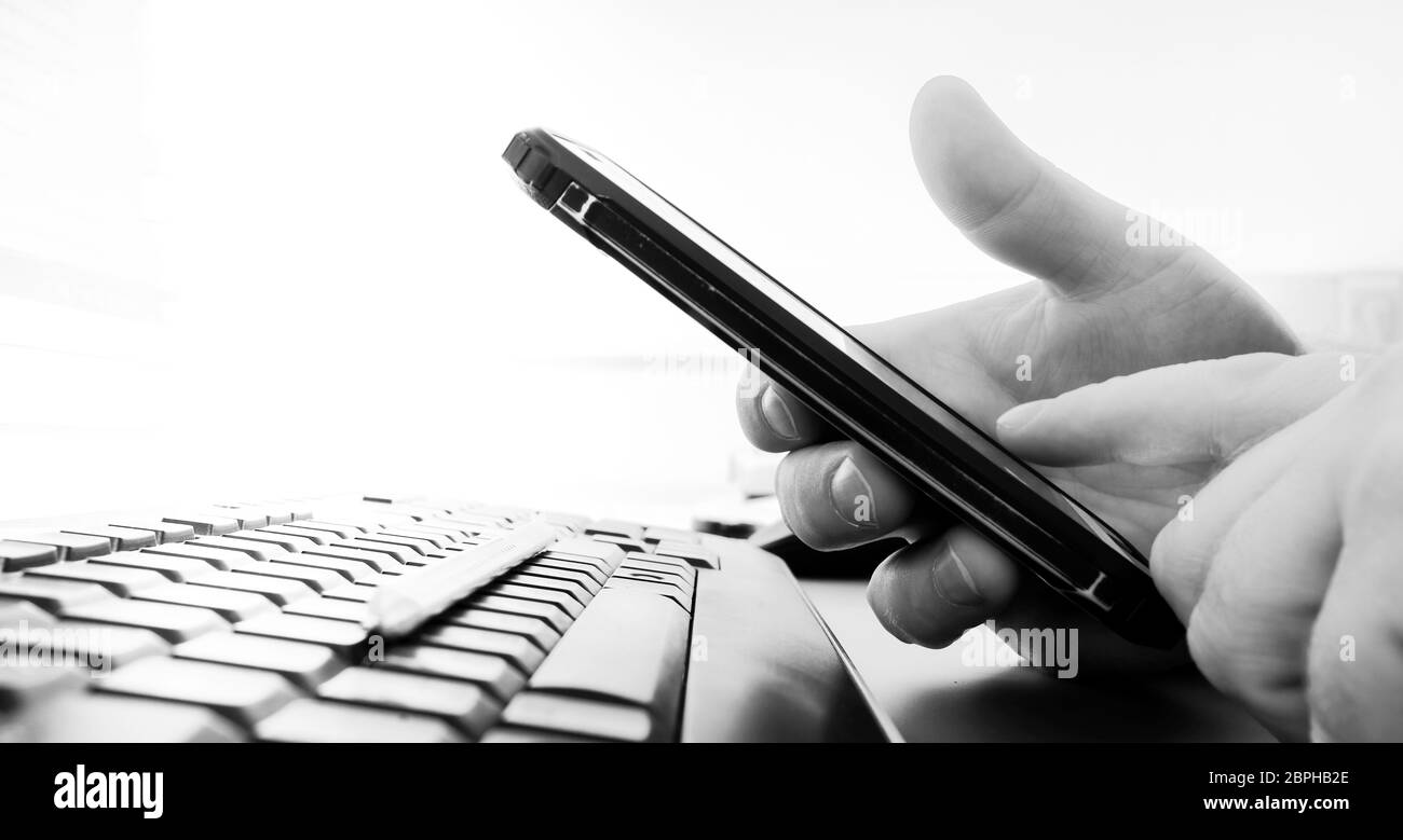 La main de l'homme tient un smartphone sur un clavier d'ordinateur noir. Arrière-plan fortement flou. Image en noir et blanc. Copier l'espace. Banque D'Images