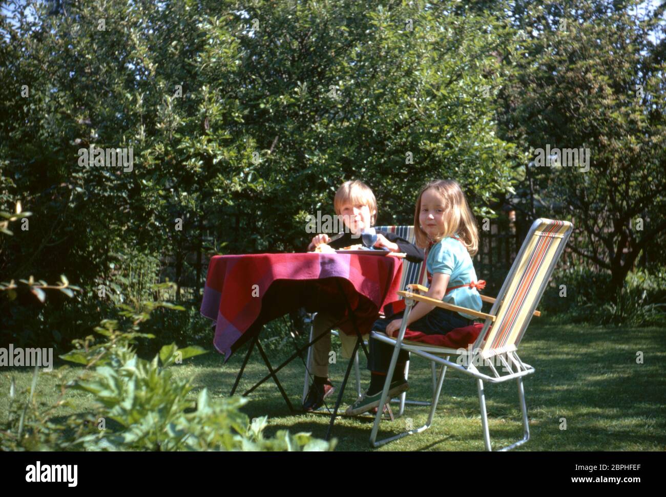 Un jeune frère et une sœur souriant, assis à une table pour prendre le petit déjeuner dans le jardin arrière de leur maison familiale dans la banlieue, Hatch End Park Estate, Milne Feild, Hatch End, Middlesex, Royaume-Uni Banque D'Images