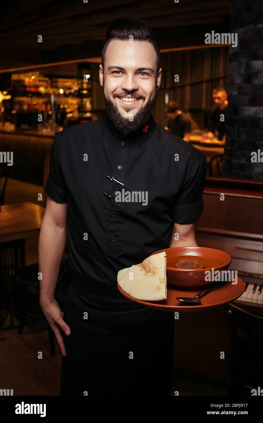 Serveur à barbe gai avec soupe de tomates riche, bœuf, riz. Un serveur éclairé en vêtements noirs, avec une barbe et une soupe de tomates Banque D'Images