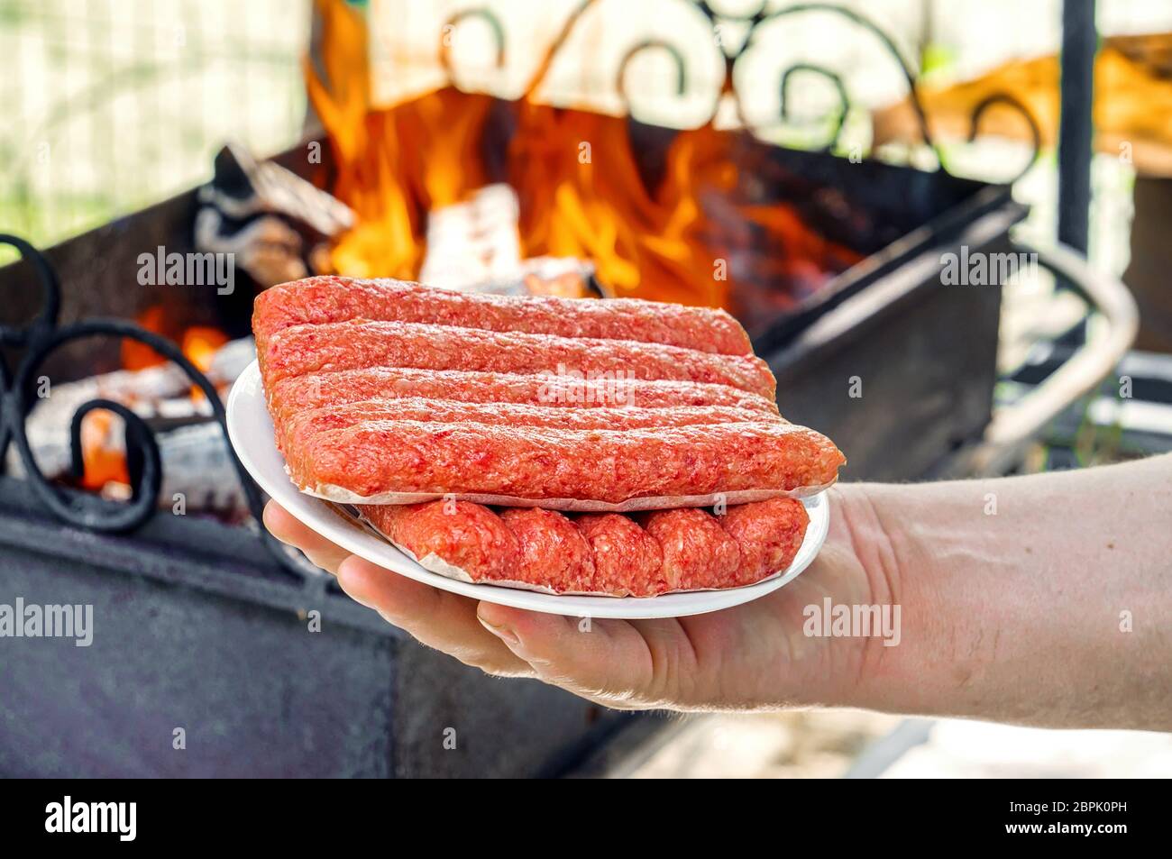 Chevapchichi sur une assiette dans votre main. Un homme tient le chevechichi frais sur un plateau blanc sur le fond d'un feu de barbecue. Chevechich grillé Banque D'Images