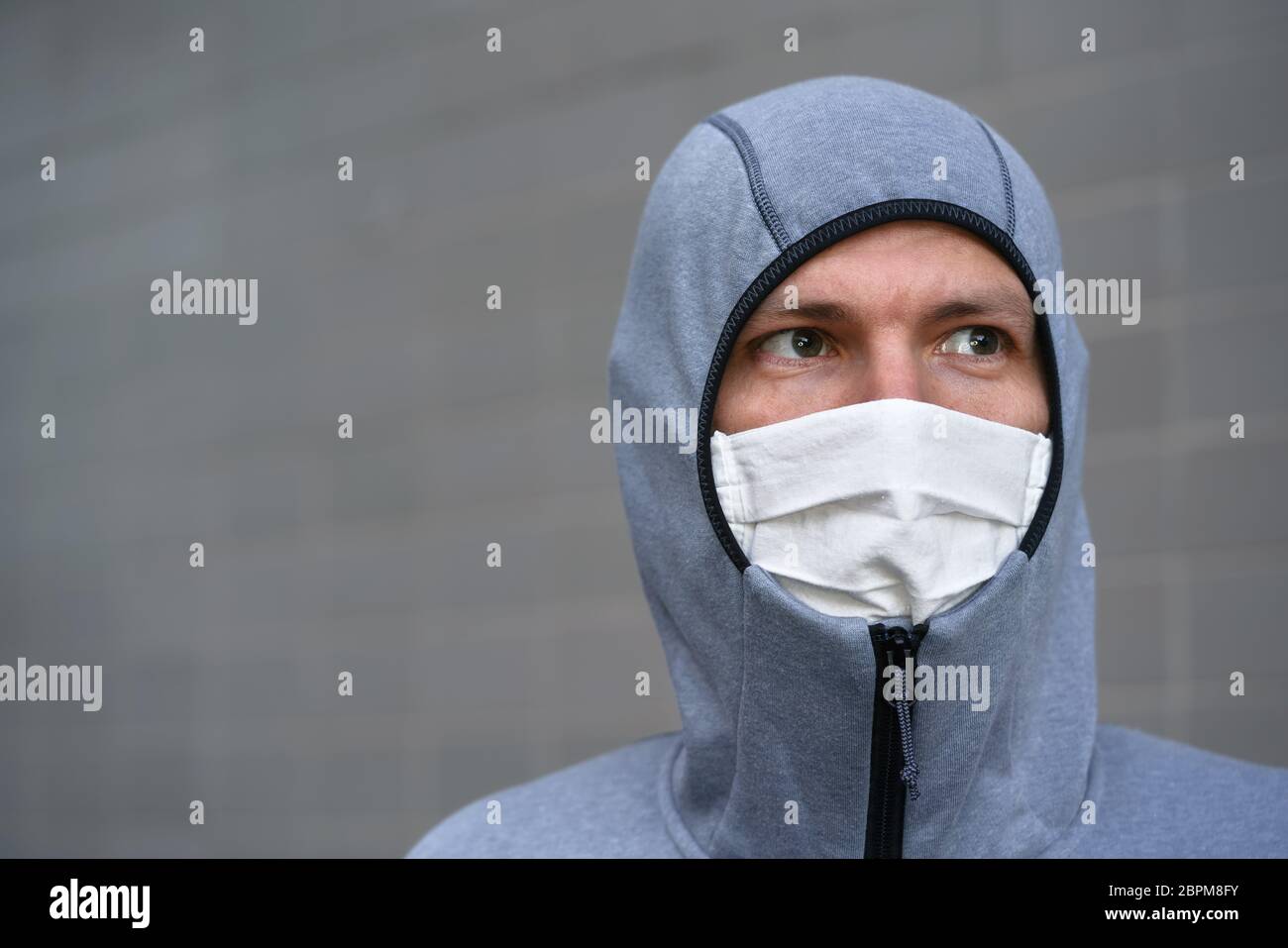 Portrait du jeune homme en sweat à capuche gris zippé et en coton fait maison visage bouche virus masque, seulement les yeux visibles, mur gris en arrière-plan. Coronavirus covid- Banque D'Images