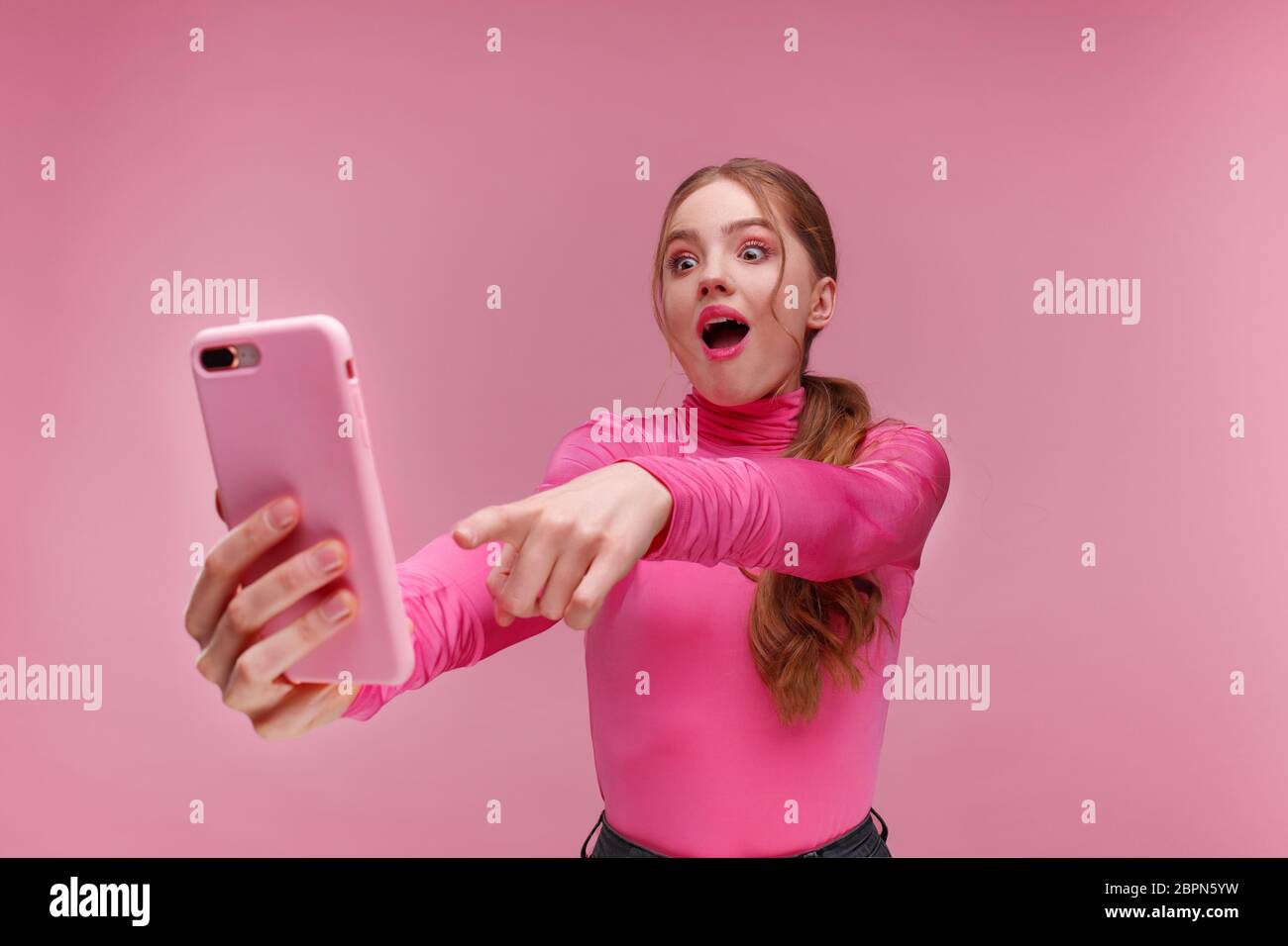 Le meilleur jour de ma vie. Surpised jeune femme rouge tenant un smartphone rose, souriant et exprimant la positivité. Une fille heureuse a reçu des nouvelles positives choquantes Banque D'Images