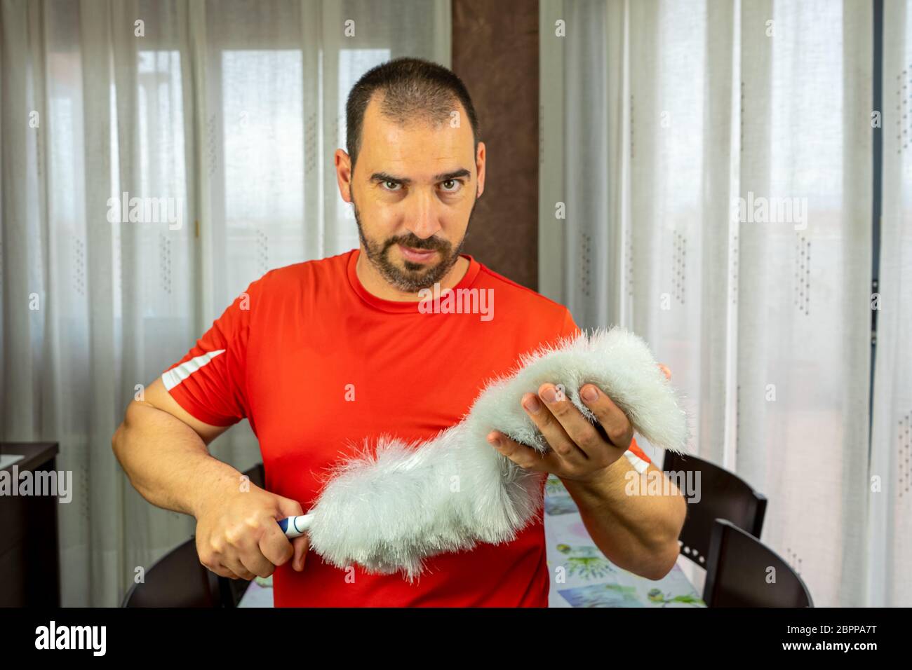 Homme avec une barbe et des cheveux courts dans une chemise rouge avec un plumeau pour nettoyer le poteau dans le salon à la maison. Concept de nettoyage Banque D'Images