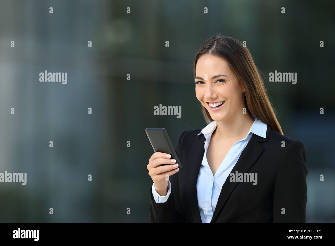 Cadre heureux utilisant un smartphone et regardant la caméra debout dans la rue avec un immeuble de bureaux en arrière-plan Banque D'Images
