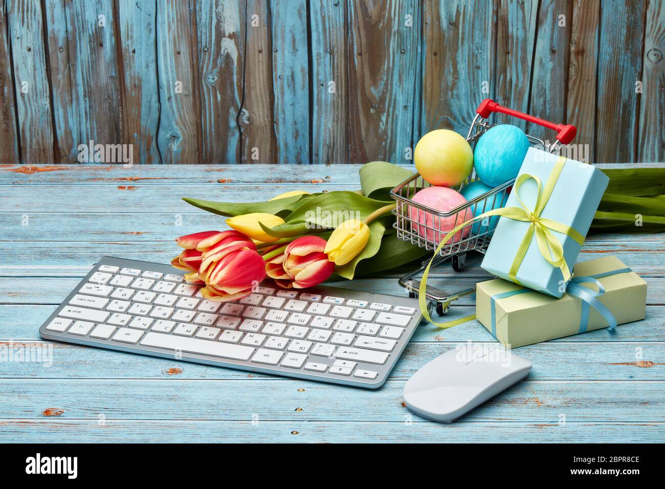 Œufs de pâques colorés dans un chariot à provisions, boîtes cadeaux, tulipes fleurs et clavier et souris d'ordinateur sur un fond en bois avec espace de copie. Pâques Banque D'Images
