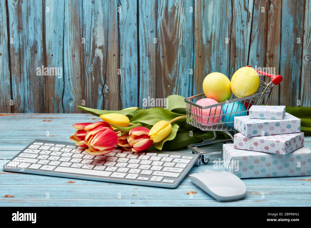 Œufs de pâques colorés dans un chariot à provisions, boîtes cadeaux, tulipes fleurs et clavier et souris d'ordinateur sur un fond en bois avec espace de copie. Pâques Banque D'Images