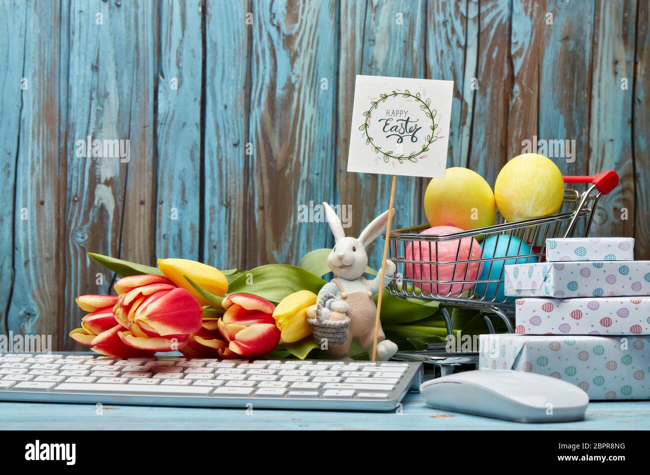 Oeufs de pâques colorés dans un chariot de shopping, boîtes-cadeaux, lapin de pâques, fleurs de tulipes et clavier d'ordinateur et souris sur un fond de bois avec copie Banque D'Images