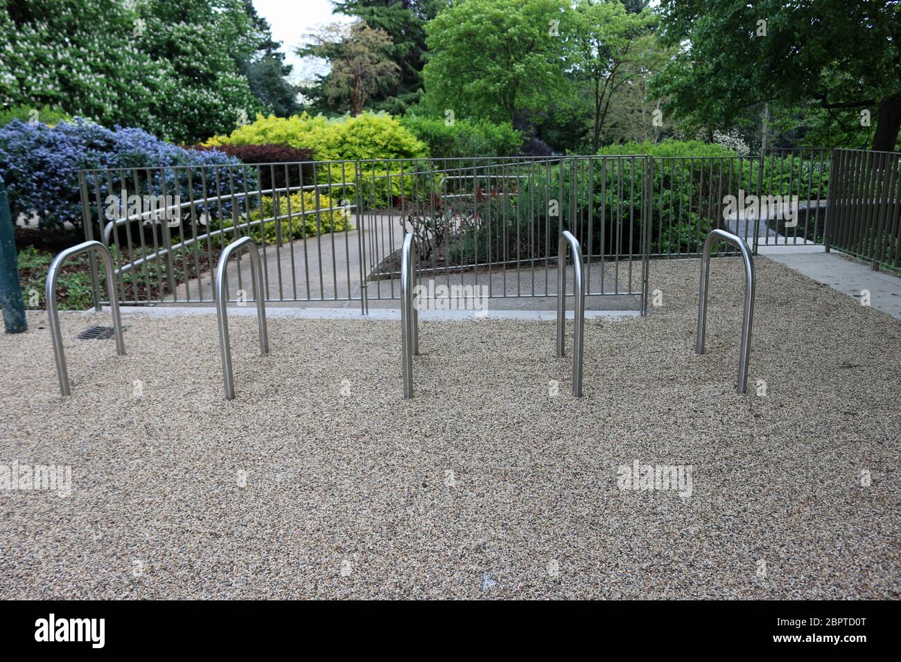 Cinq nouveaux métal des supports à vélo sur une piste en gravier chemin dans un parc avec un arbre, des lits d'arbustes, de clôtures et d'autres équipement de parc derrière. Banque D'Images