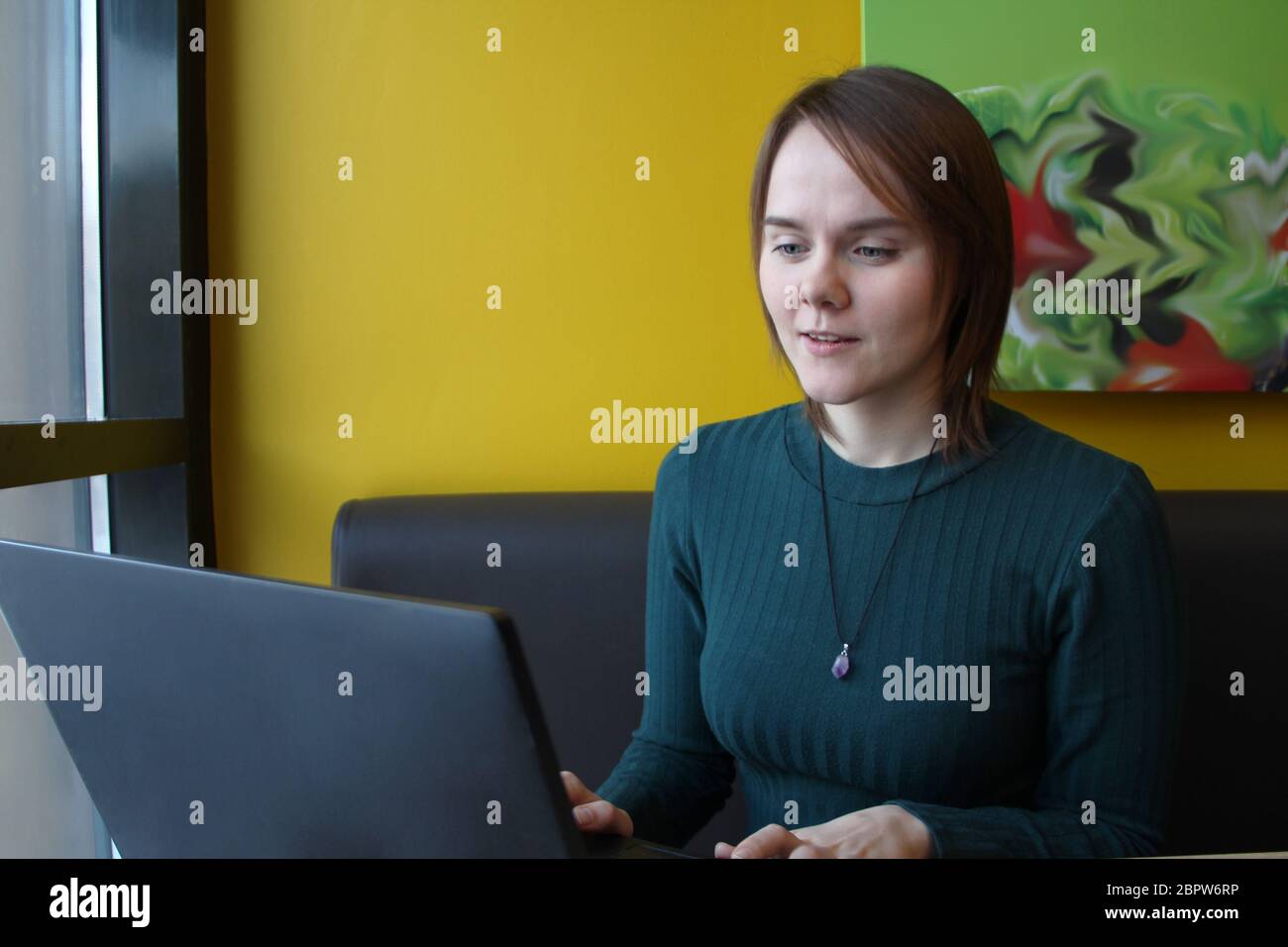 Belle jeune fille assise dans un café à une table avec un ordinateur portable. Fonctionne sur l'ordinateur et saisit sur le clavier. Regarde l'écran avec une expression émotionnelle. Banque D'Images