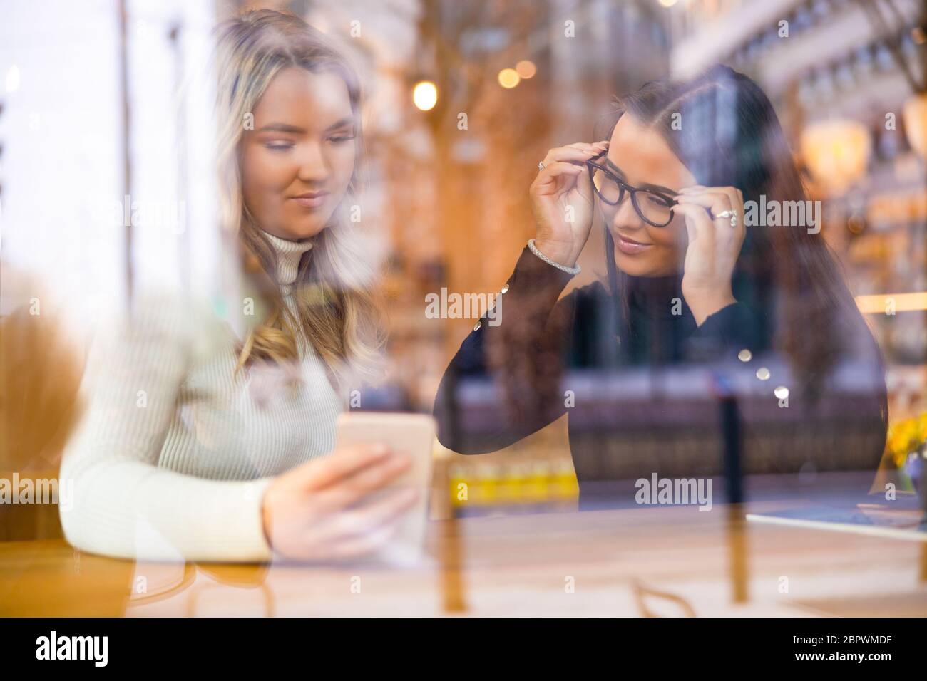 Deux belles femmes regardant le téléphone ensemble dans le café vu par la fenêtre Banque D'Images