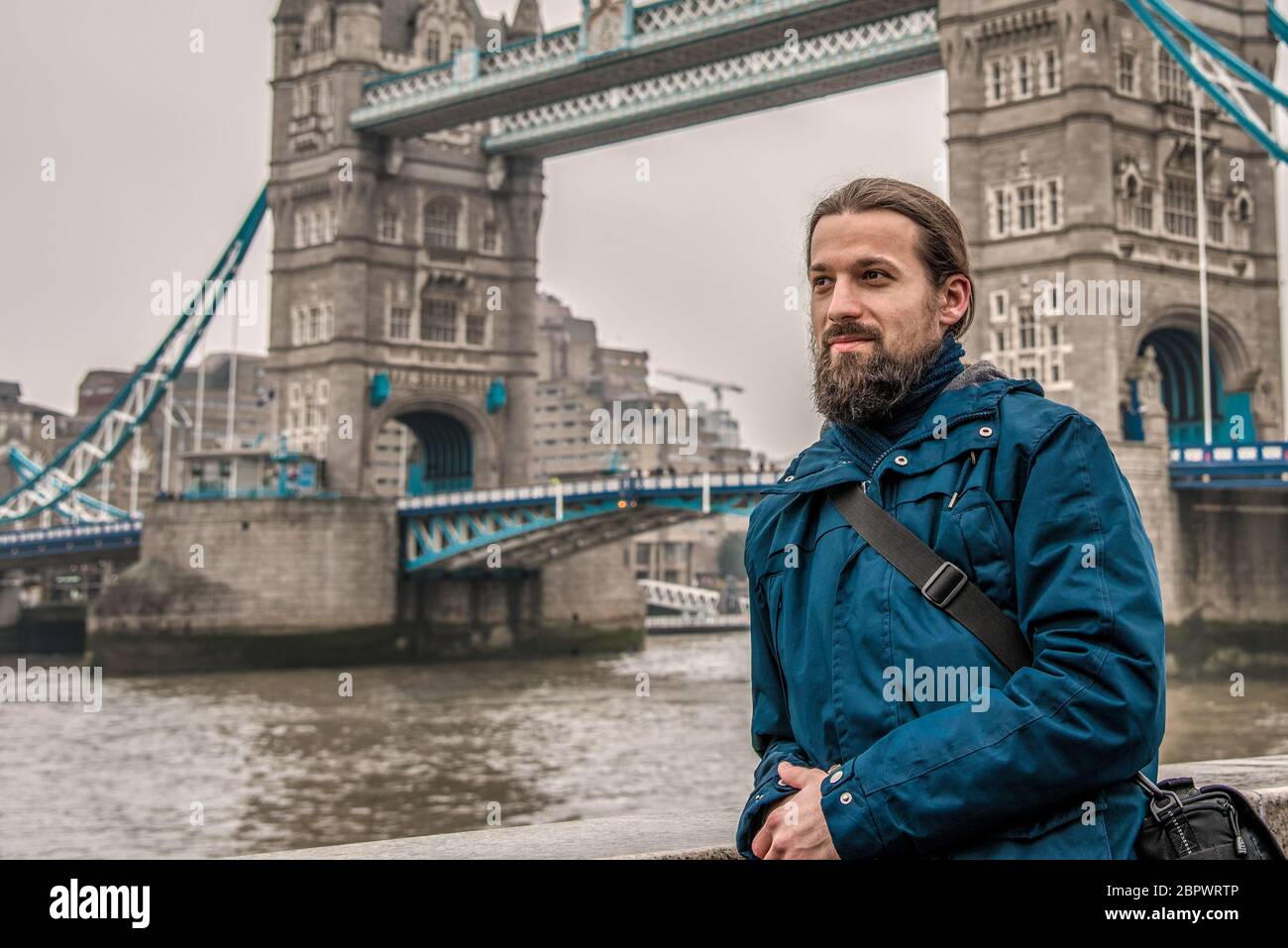 Un jeune touriste mâle aux cheveux longs et à la barbe épaisse se tient sur le fond du London Bridge en hiver par temps nuageux Banque D'Images