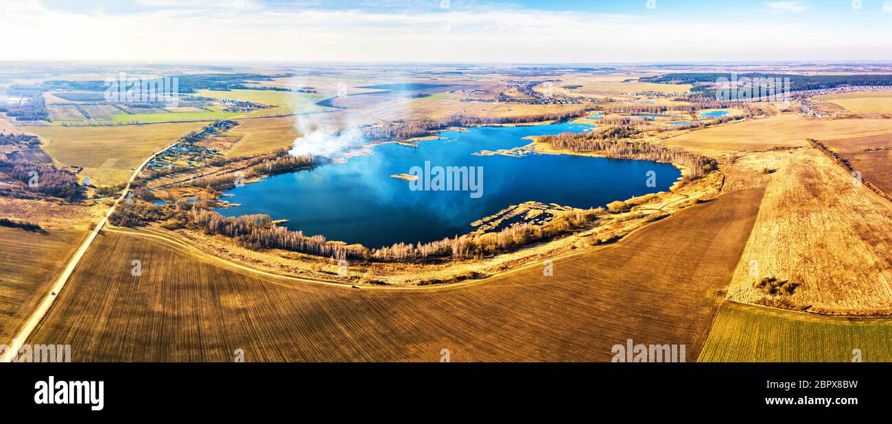 Printemps des champs agricoles. De la fumée sur l'eau et le lac bleu panorama. Les cultures d'hiver et champ labouré. Campagne de printemps paysage aérien. Des chemins de terre Banque D'Images