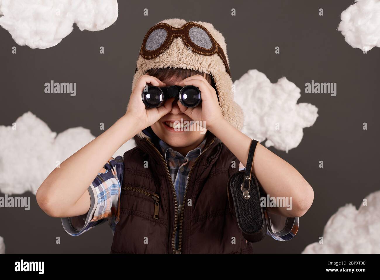 un garçon joue avec des jumelles et rêve de devenir pilote, habillé dans une veste rétro et un casque avec des lunettes, des nuages de laine de coton, un dos gris Banque D'Images
