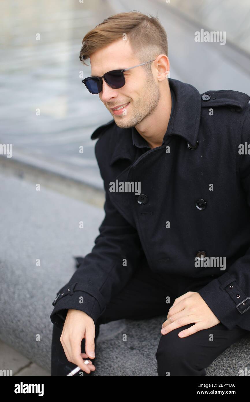 Jeune homme blond souriant portant une veste noire assis sur la Pyramide du Louvre et fumant. Banque D'Images