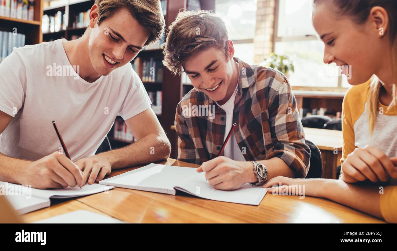 Groupe d'élèves assis à la table dans la bibliothèque et étudiant ensemble. De jeunes amis heureux travaillant sur le projet de lycée. Banque D'Images
