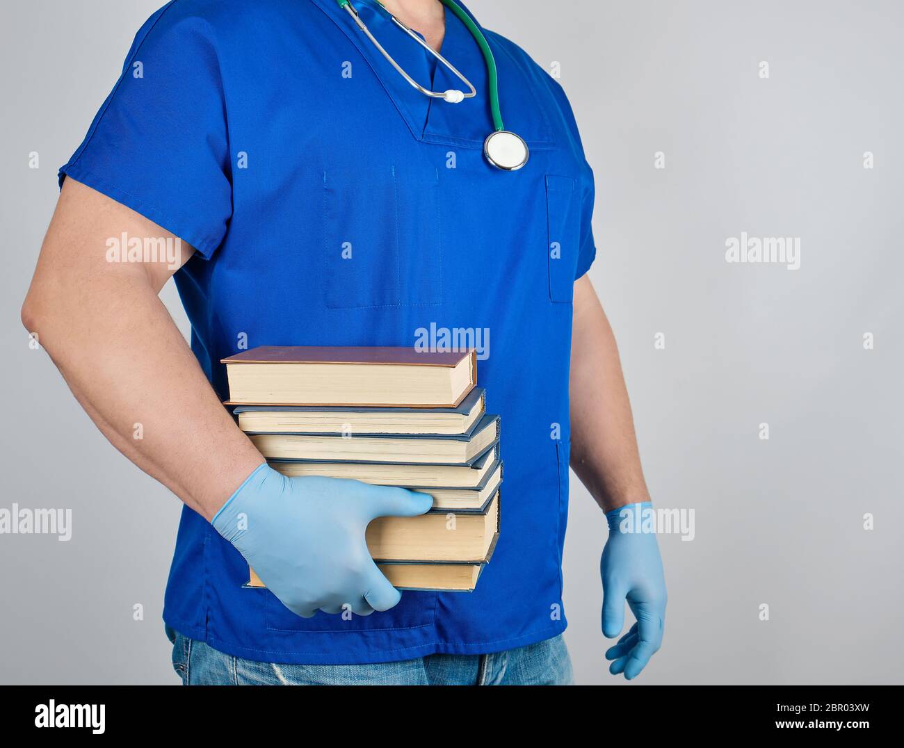 Médecin en uniforme bleu et des gants latex stériles tient dans sa main une pile de livres sur un fond gris, l'homme se tient sur le côté Banque D'Images