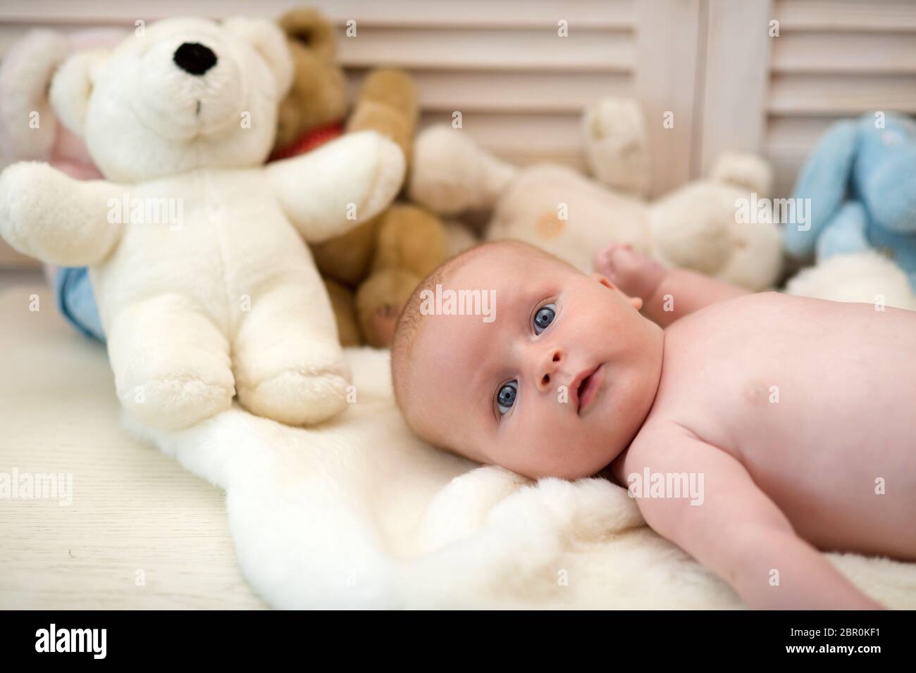 Bébé avec les yeux bleus et le visage surpris sur une couverture légère  avec des jouets sur fond, défoqué. Bébé couché sur une couette blanche. Bébé  garçon et son ours en peluche