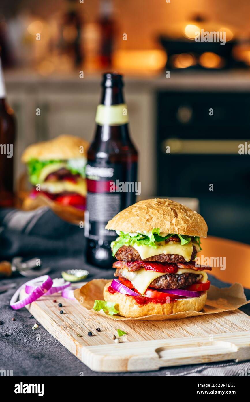Cheeseburger avec deux galettes de boeuf, fromage cheddar, bacon, laitue Iceberg, des tranches de tomate et d'oignon rouge. Bouteille de bière et des ingrédients sur tabl Banque D'Images