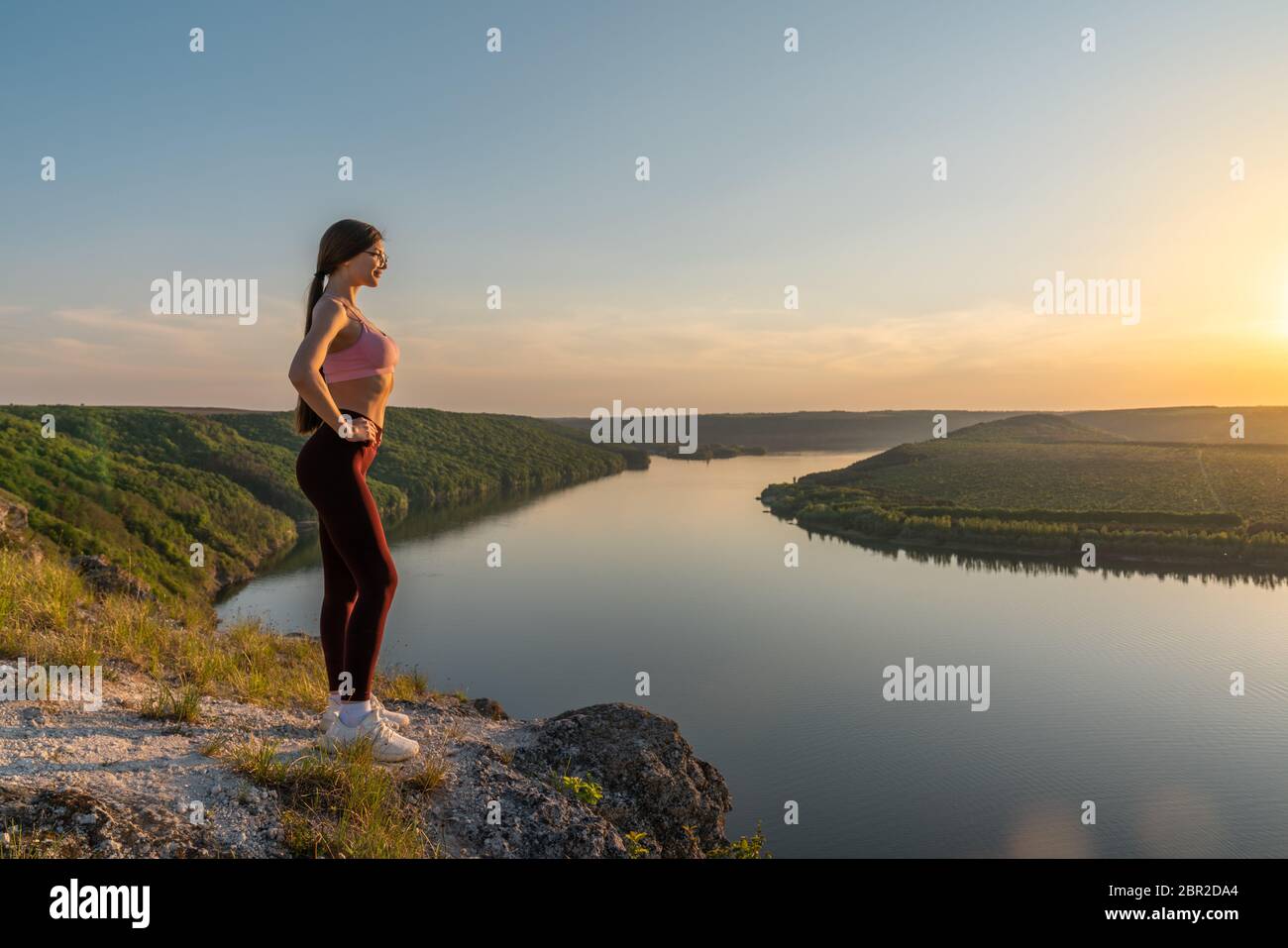 Jeune femme active de randonneur dans des vêtements de sport et des lunettes se détendant sur un rocher à haute altitude, regardant le magnifique panorama avec le coucher du soleil Banque D'Images