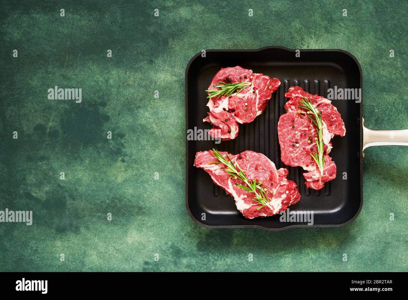 Trois steaks de bœuf frais, crus, au romarin et au poivre, dans une casserole sur fond de béton vert. Vue de dessus, espace de copie pour le texte. Banque D'Images