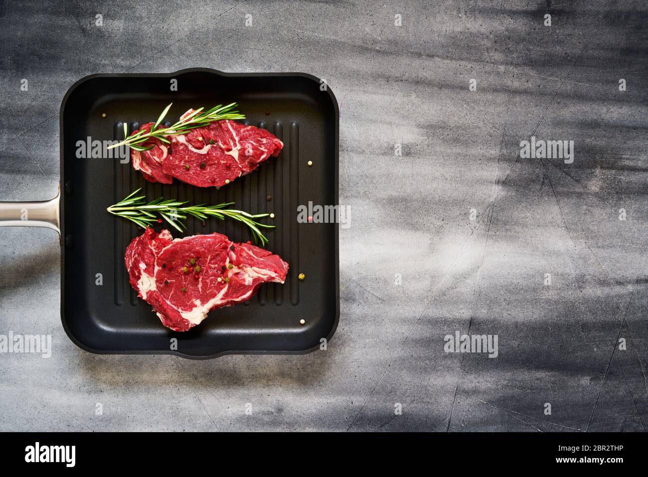 Deux steaks de bœuf frais, crus, au romarin et au poivre, dans une casserole sur fond de béton gris. Vue de dessus, espace de copie pour le texte. Banque D'Images