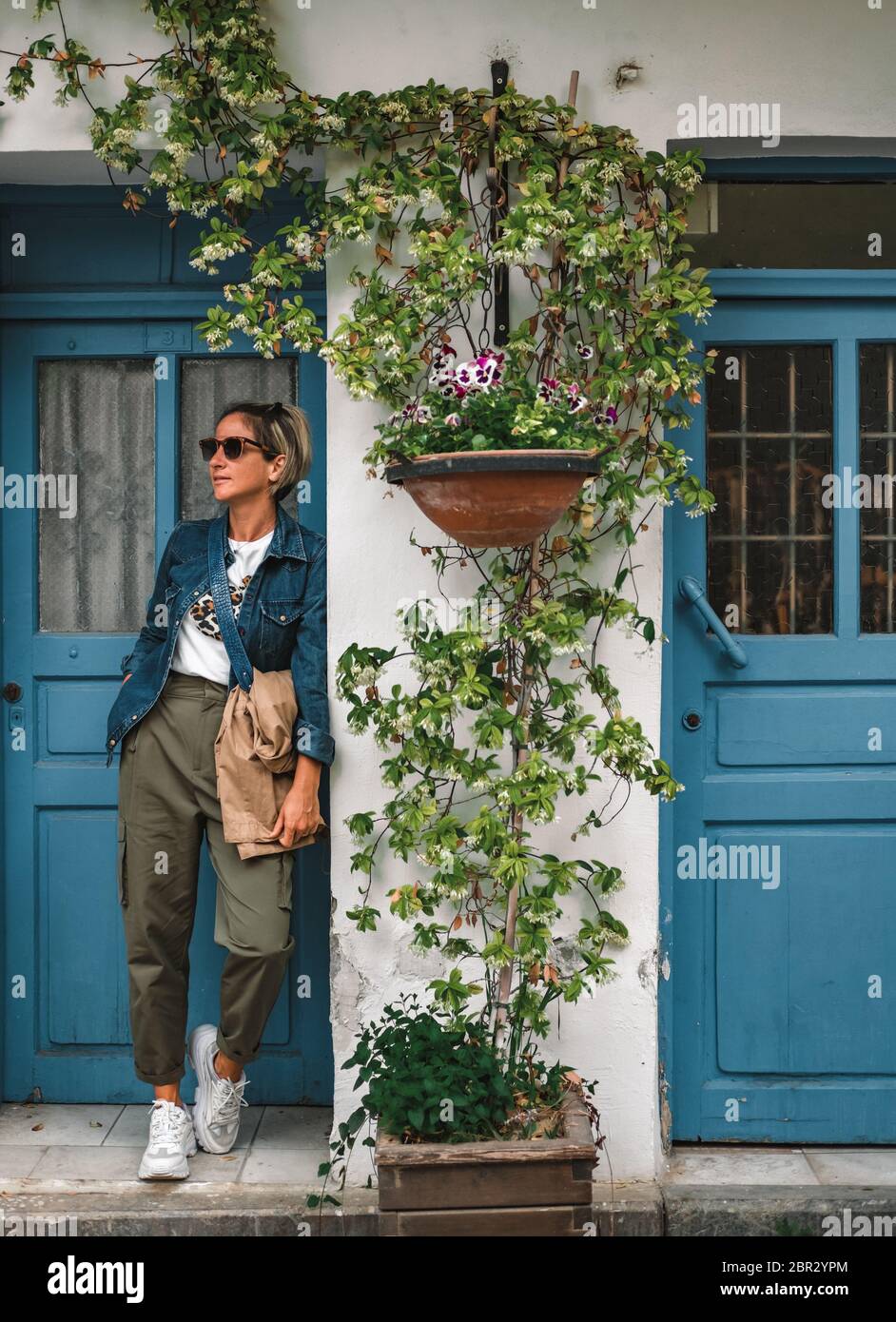 Jolie femme à la mode debout dans la porte de la vieille maison. Dame fraîche et habillée devant une porte en bois turquoise avec des fleurs. Banque D'Images Jolie femme à la mode debout dans la porte de la vieille maison. Dame fraîche et habillée devant une porte en bois turquoise avec des fleurs. Banque D'Images