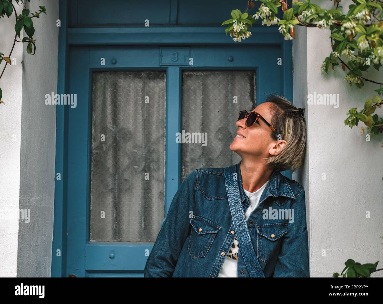 Jolie femme à la mode debout dans la porte de la vieille maison. Dame fraîche et habillée devant une porte en bois turquoise avec des fleurs. Banque D'Images Jolie femme à la mode debout dans la porte de la vieille maison. Dame fraîche et habillée devant une porte en bois turquoise avec des fleurs. Banque D'Images