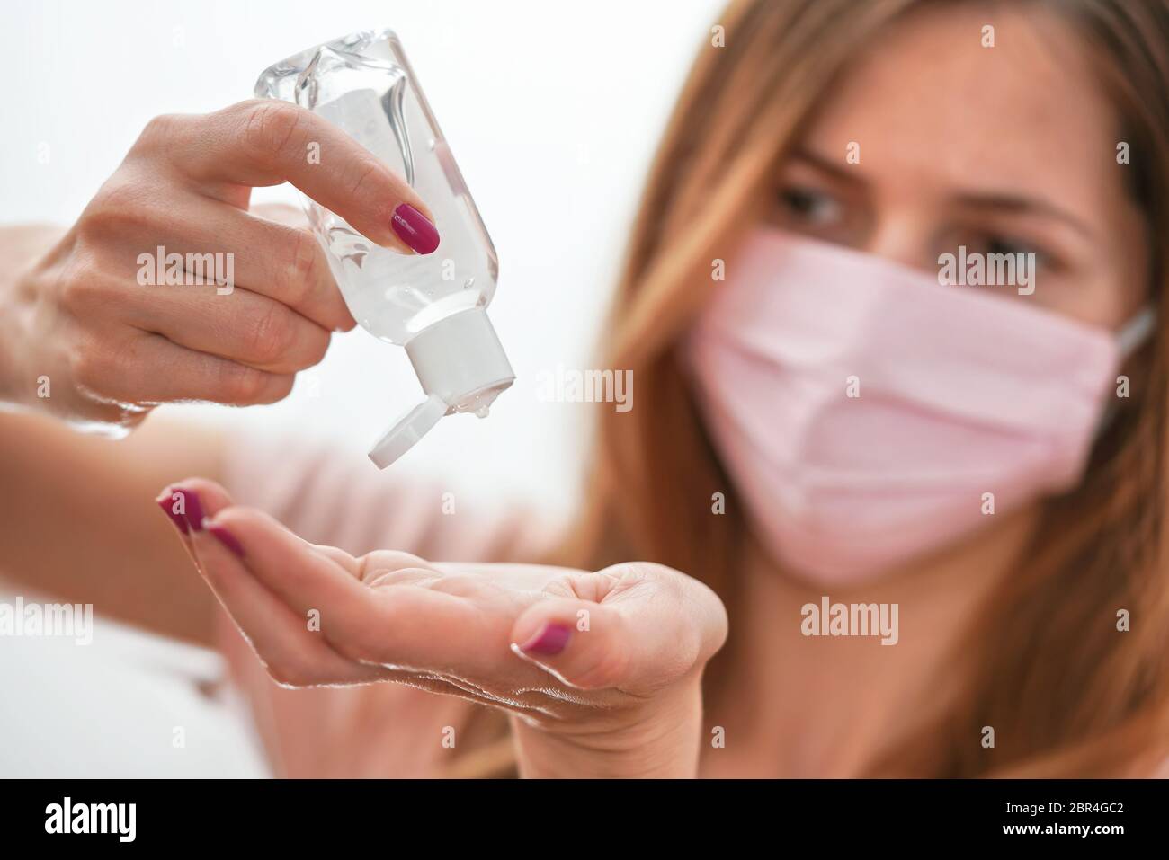 Jeune femme avec du coton rose fait maison bouche visage virus masque compte-gouttes anti bactérien alcool gel à frotter sur la main, détail à la bouteille blanche, visage flou Banque D'Images