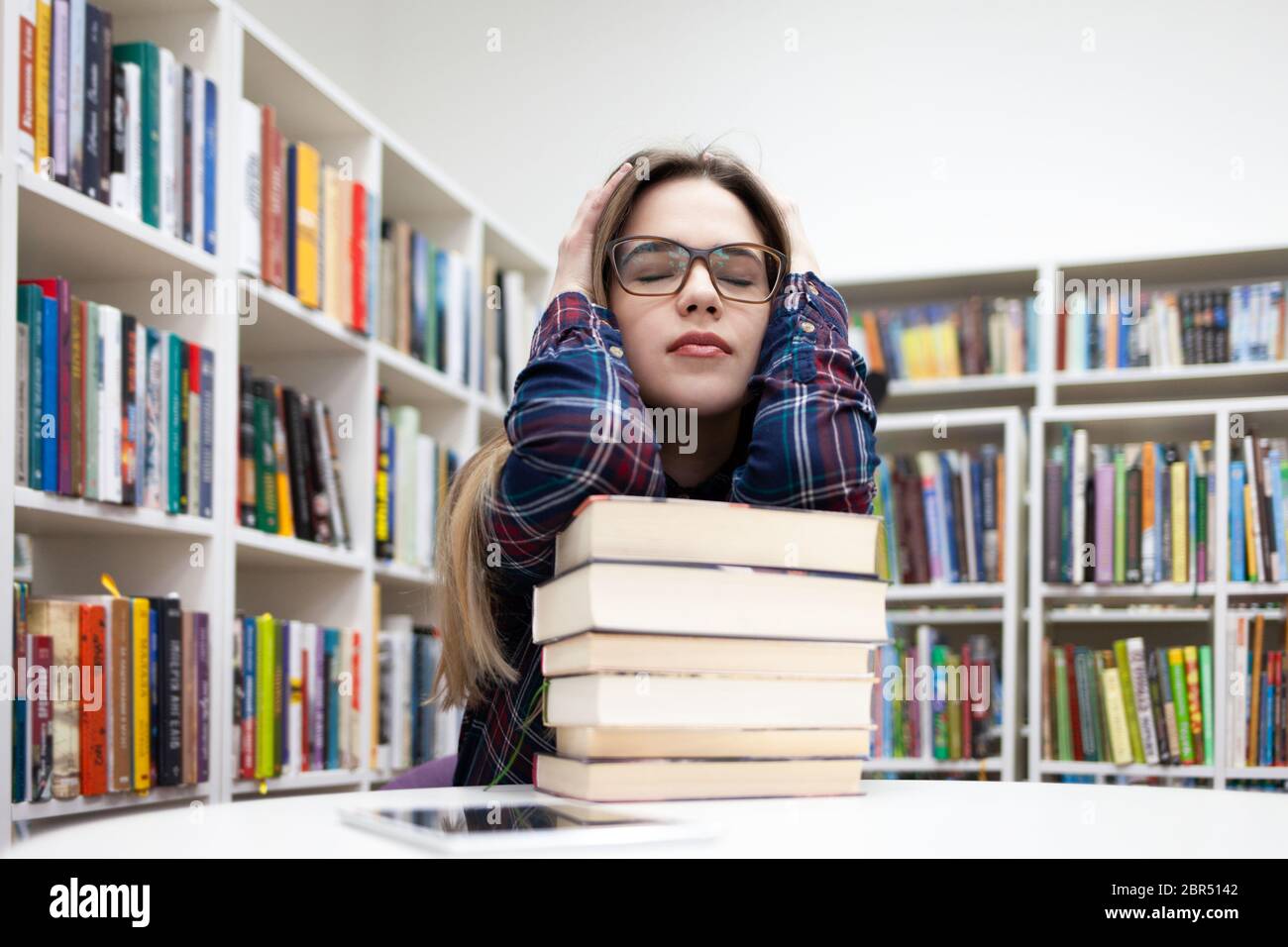 Jeune fille étudiante fatiguée assise dans une bibliothèque à une table  penchée sur une grosse pile de livres. La femme dépressive dans une chemise  à carreaux se prépare pour les examens dans