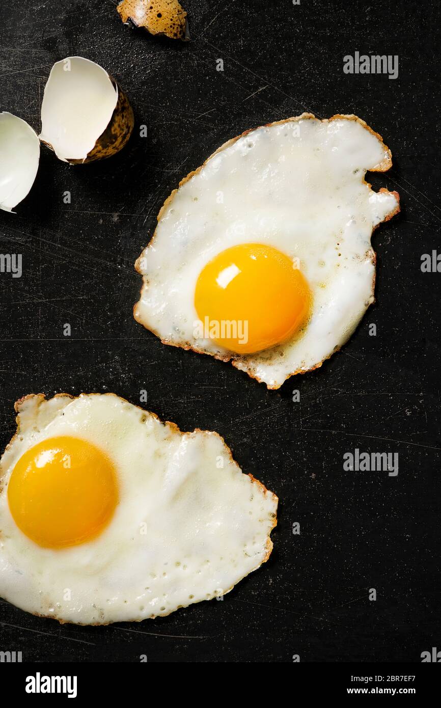 2 œufs de caille frits sur la surface noire de la table de cuisson avec des coquilles d'œufs fissurées sur le côté. Banque D'Images